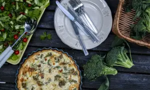 En paj med ost och broccolisås, en sallad och färsk broccoli ligger uppdukat på ett träbord.