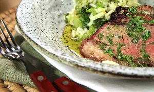 Close-up of grilled picanha steak with rice and greens, served on a speckled plate with a fork and knife.