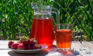 En karaff och ett glas med röd saft står på ett träbord. En tallrik med färska jordgubbar bredvid. Perfekt för ett enkelt jordgubbssaft recept.