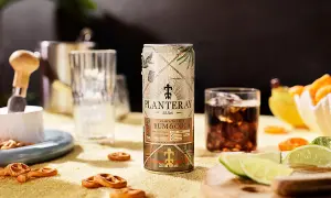 En burk Planteray Rum & Cola står på ett gult underlag, bredvid ett glas med is och mörk dryck. Små pretzels och limeklyftor ligger utspridda.