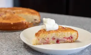 En bit saftig sockerkaka med rabarber, toppad med en klick vispgrädde. Hela kakan syns i bakgrunden.