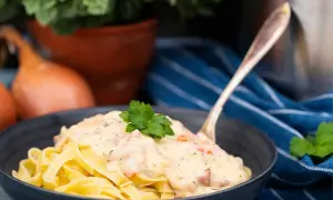 En skål med tagliatelle täckt av krämig ost och skinksås, toppad med persilja. En gaffel sticker upp ur pastan.