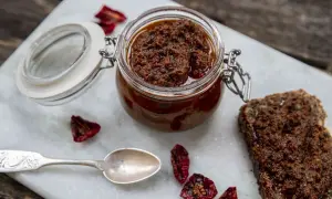 En burk med soltorkad tomat-tapenade, perfekt för ett enkelt tapenade recept. Serveras med en skiva mörkt bröd och en silver sked.