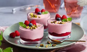 Tre glas med hallonmousse tårta, toppade med hallon, blåbär och pistagenötter. Serveras på en ljusgrön tallrik.