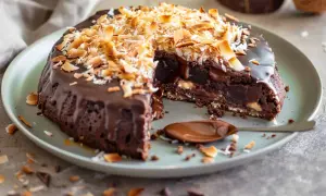 En saftig kladdkaka med chokladglasyr och rostad kokostosca. En bit saknas, och en sked med chokladsås ligger bredvid.