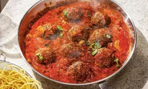 Frikadeller i tomatsås som puttrar i en panna, med en skål spaghetti vid sidan.