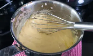 En visp rör i en gul sås i en kastrull. Små bitar av schalottenlök syns på kastrullkanten. Ett perfekt bearnaisesås recept börjar här.