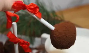 En hand håller en chokladboll på pinne med röd rosett, redo att doppas i en mugg med varm choklad.