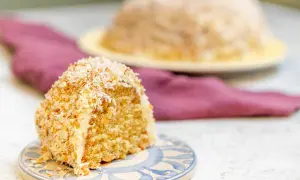 En bit sockerkaka med kokos, täckt av rostad kokosflingor. En hel kaka syns i bakgrunden.