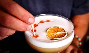 En hand dekorerar en whiskey sour med en tunn pinne, med en torkad lime och ett mönster av bitters på skummet.