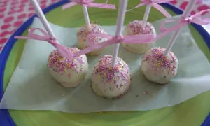 Sju vita cake pops med rosa och gula strössel, dekorerade med rosa band, ligger på ett ljusgrönt papper på en grönblå tallrik.