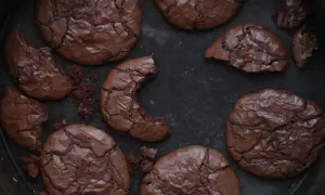 Mörka, spröda nutella kakor med sprucken yta ligger utspridda i en form. En kaka är avbruten och visar det saftiga inre.