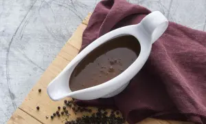 En vit såssip med mustig brunsås, redo för ett klassiskt brunsås recept. Svartpepparkorn och en vinröd duk fulländar bilden.