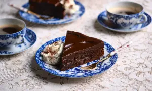 En bit kladdkaka med chokladganache och vispad grädde, serverad med kaffe på blåvita koppar.