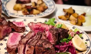 En saftig bistecca fiorentina, perfekt grillad med en rosa kärna, serveras med krispig sallad och citronklyfta. Rostade potatisar i bakgrunden.