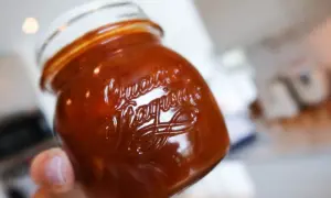 En hand håller en burk med hemlagad rabarberchutney. Den mörkröda chutneyn glänser i ljuset.