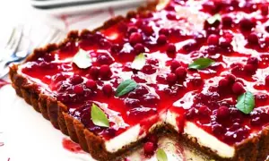 En rektangulär pepparkakscheesecake med en glansig lingonfyllning och färska lingon. Garnerad med gröna blad.