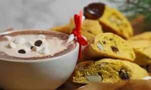 En kopp varm choklad med marshmallows och chokladbitar bredvid en hög med saffransbiscotti med mandel, inslagna i ett rött band.