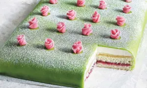 En grön täckt liten tårta med rosa marsipanrosor och pudersocker. En bit är bortskuren och visar lager av grädde och sylt.