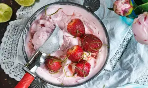 En skål med rosa jordgubbsglass, toppad med hela jordgubbar och limezest. En glasskopa vilar i skålen.