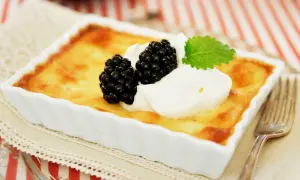 En gyllene, gammaldags ostkaka recept i en vit form, toppad med vispgrädde, björnbär och en myntakvist. Serveras med en gaffel.