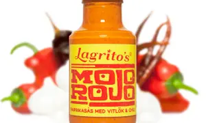 En flaska Lagrito's Mojo Rojo paprikasås med vitlök och chili, omgiven av suddiga röda chilifrukter och vitlöksklyftor.