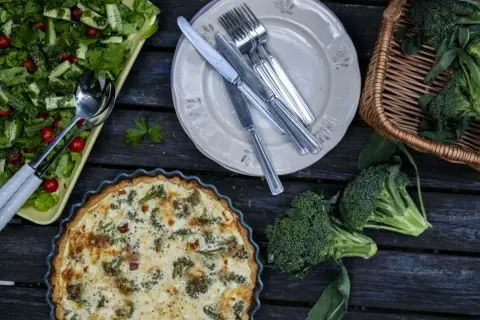 En paj med ost och broccolisås, en sallad och färsk broccoli ligger uppdukat på ett träbord.