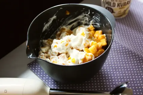 En skål med fruktbitar och krämig kvarg, redo att bli en härlig kvarg glass. En stavmixer ligger bredvid.