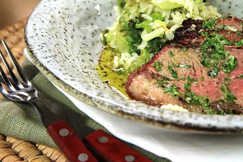 Close-up of grilled picanha steak with rice and greens, served on a speckled plate with a fork and knife.
