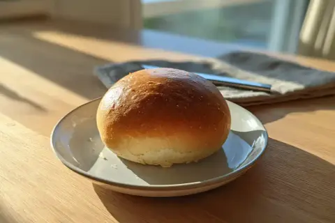En gyllene, luftig semla på en tallrik, redo att fyllas med grädde och mandelmassa. Solen lyser in genom fönstret.