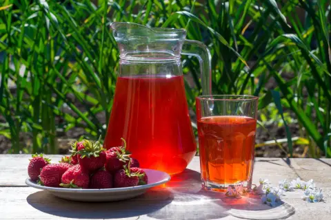 En karaff och ett glas med röd saft står på ett träbord. En tallrik med färska jordgubbar bredvid. Perfekt för ett enkelt jordgubbssaft recept.