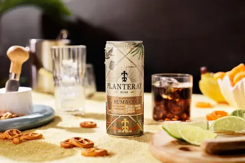 En burk Planteray Rum & Cola står på ett gult underlag, bredvid ett glas med is och mörk dryck. Små pretzels och limeklyftor ligger utspridda.