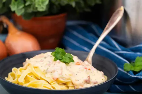 En skål med tagliatelle täckt av krämig ost och skinksås, toppad med persilja. En gaffel sticker upp ur pastan.