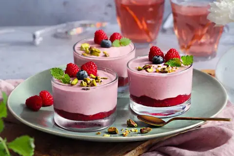 Tre glas med hallonmousse tårta, toppade med hallon, blåbär och pistagenötter. Serveras på en ljusgrön tallrik.