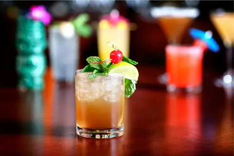 En Mai Thai-drink med is, mynta, lime och körsbär. I bakgrunden syns fler drinkar och en grön tiki-mugg.