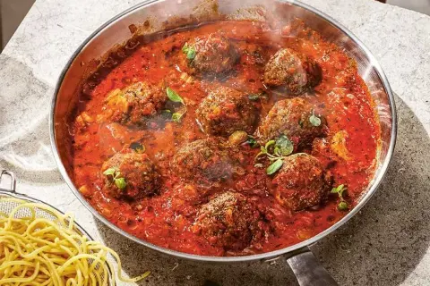 Frikadeller i tomatsås som puttrar i en panna, med en skål spaghetti vid sidan.