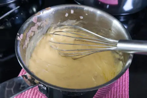 En visp rör i en gul sås i en kastrull. Små bitar av schalottenlök syns på kastrullkanten. Ett perfekt bearnaisesås recept börjar här.