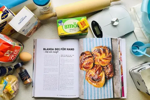 Här finns allt för ett lyckat kanelbullar recept: mjöl, jäst, smör, socker, kardemumma och kanel. En öppen bok visar hur man gör.