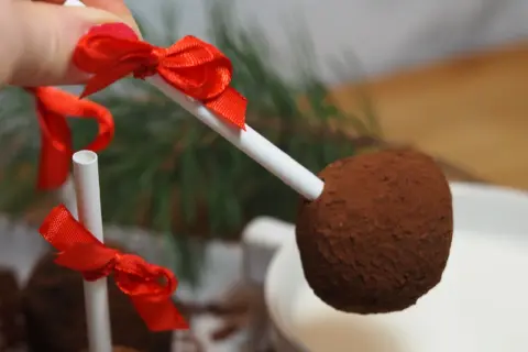 En hand håller en chokladboll på pinne med röd rosett, redo att doppas i en mugg med varm choklad.