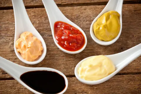 Fem skedar med olika såser: ketchup, majonnäs, senap, en orange sås och mörk bbq sås på träbakgrund.