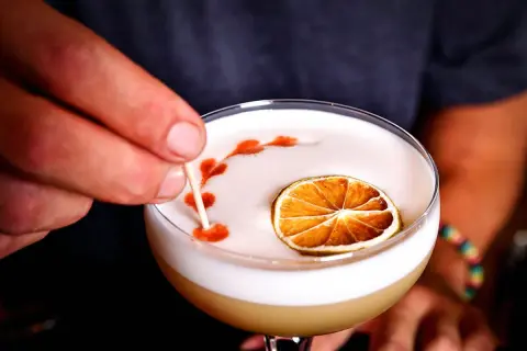 En hand dekorerar en whiskey sour med en tunn pinne, med en torkad lime och ett mönster av bitters på skummet.