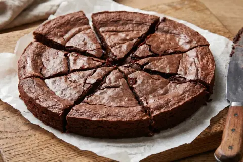 En saftig glutenfri kladdkaka, uppskuren i åtta bitar på bakplåtspapper. En kniv ligger bredvid.