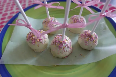 Sju vita cake pops med rosa och gula strössel, dekorerade med rosa band, ligger på ett ljusgrönt papper på en grönblå tallrik.