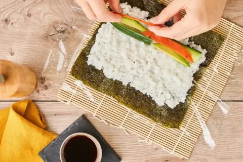 Händer lägger lax och gurka på ris för ett sushi recept.