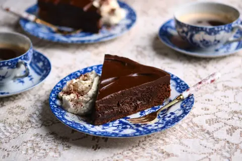 En bit kladdkaka med chokladganache och vispad grädde, serverad med kaffe på blåvita koppar.