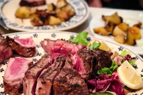 En saftig bistecca fiorentina, perfekt grillad med en rosa kärna, serveras med krispig sallad och citronklyfta. Rostade potatisar i bakgrunden.
