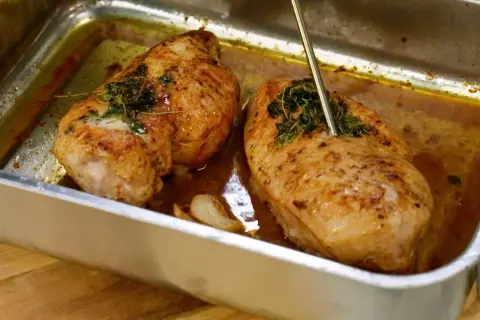 Två saftiga kycklinglårfiléer i en ugnsform, kryddade med örter och vitlök. En termometer mäter temperaturen för perfekt grillad kyckling.