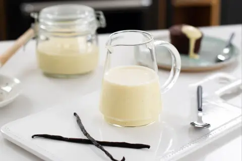 En liten kanna med krämig vaniljsås, perfekt till efterrätt. Två vaniljstänger ligger bredvid.