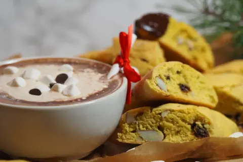 En kopp varm choklad med marshmallows och chokladbitar bredvid en hög med saffransbiscotti med mandel, inslagna i ett rött band.