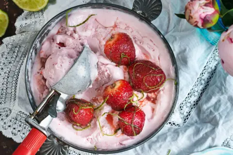 En skål med rosa jordgubbsglass, toppad med hela jordgubbar och limezest. En glasskopa vilar i skålen.